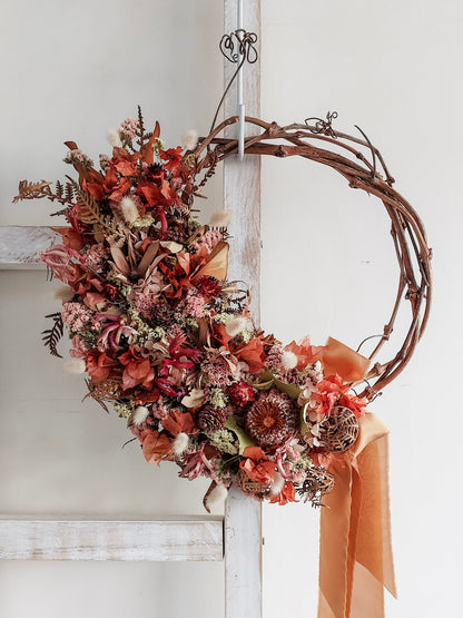 A rustic dried flower wreath featuring red, rust, and earthy dried flowers and foliage on a grapevine base, hanging on a white wooden ladder.