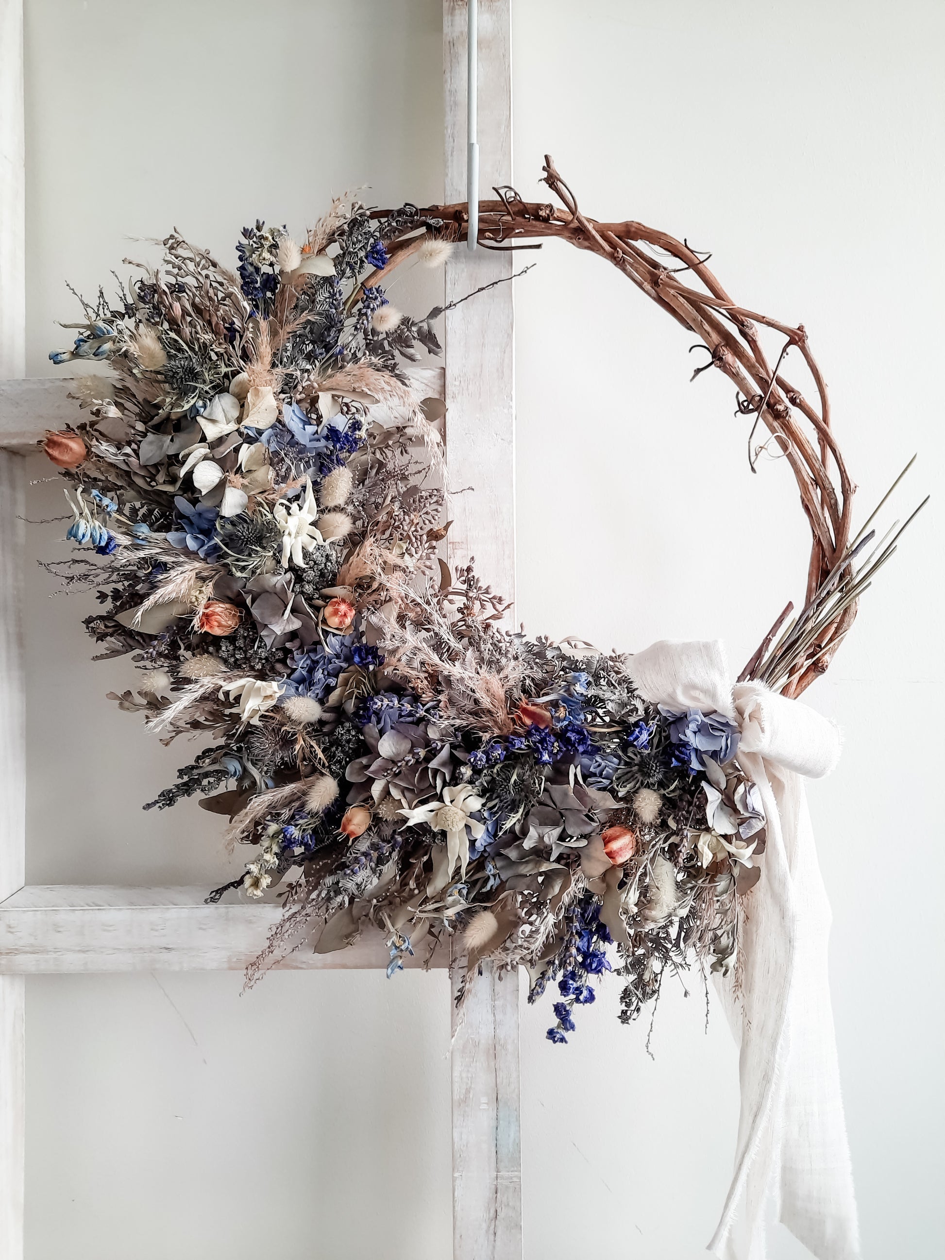 A rustic dried flower wreath featuring blue, cream, and neutral dried flowers and foliage on a grapevine base, hanging on a white wooden ladder.