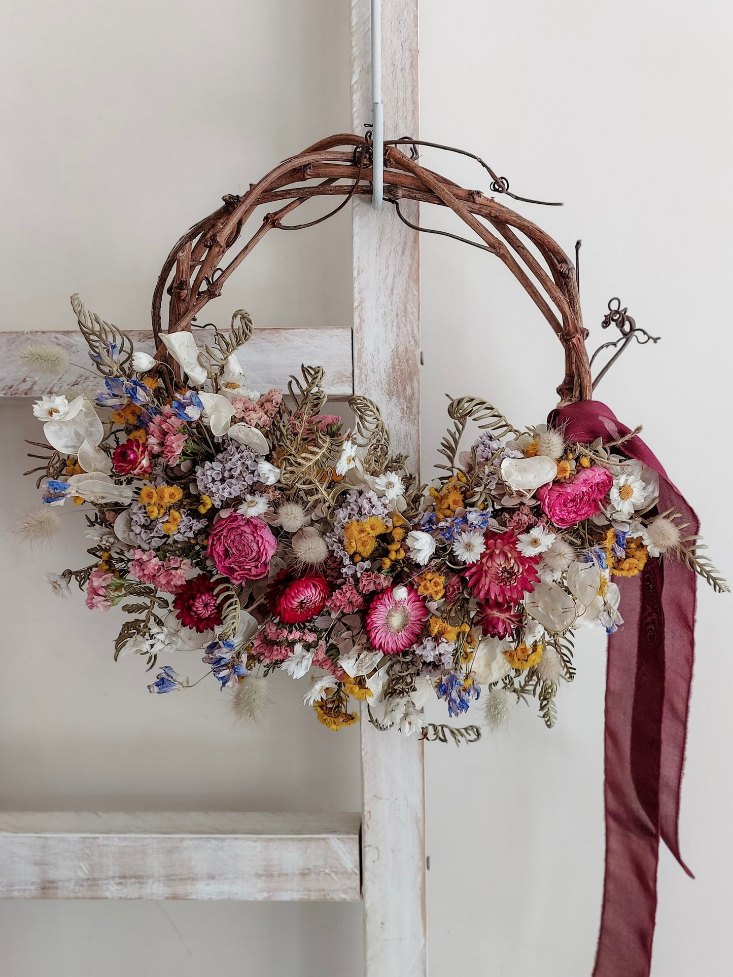 A rustic dried flower wreath featuring pink, mauve, and yellow dried flowers and foliage on a grapevine base, hanging on a white wooden ladder.