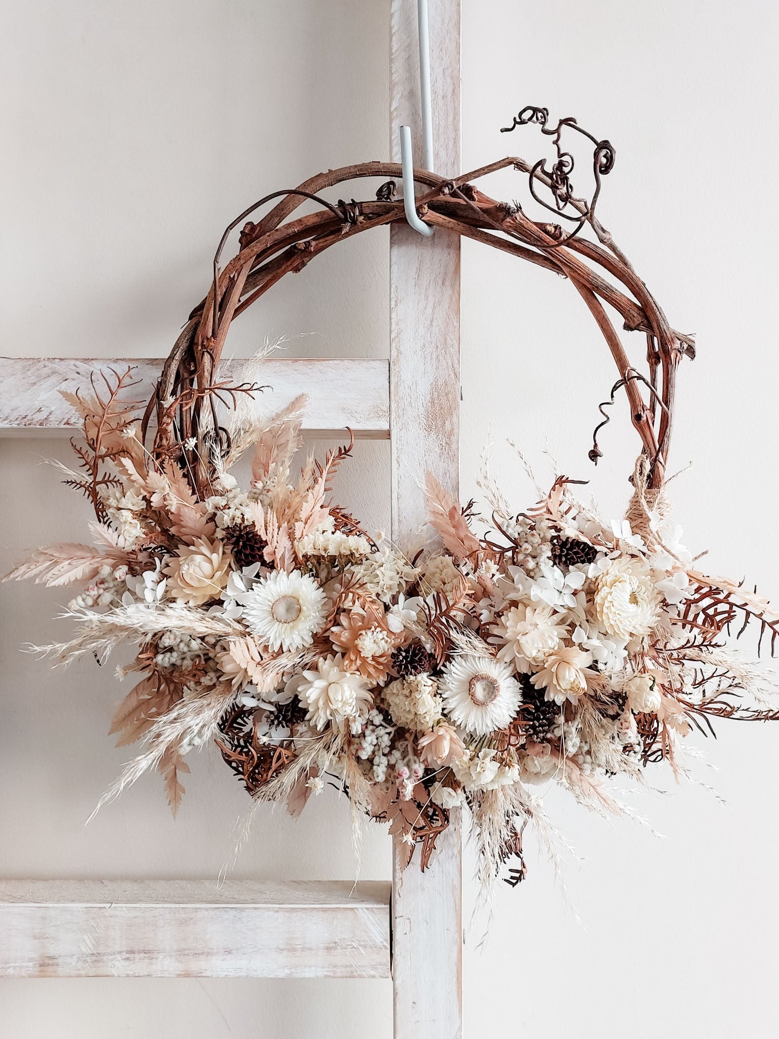 A rustic dried flower wreath featuring white, nude, and earthy dried flowers and foliage on a grapevine base, hanging on a white wooden ladder.