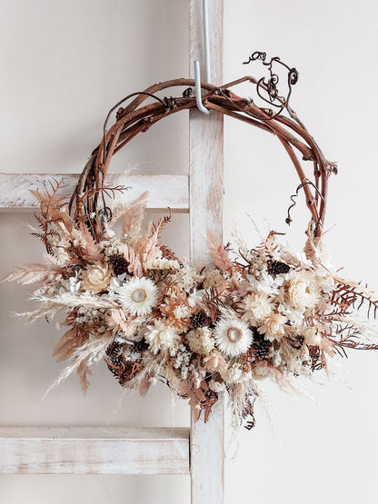 A rustic dried flower wreath featuring white, nude, and earthy dried flowers and foliage on a grapevine base, hanging on a white wooden ladder.