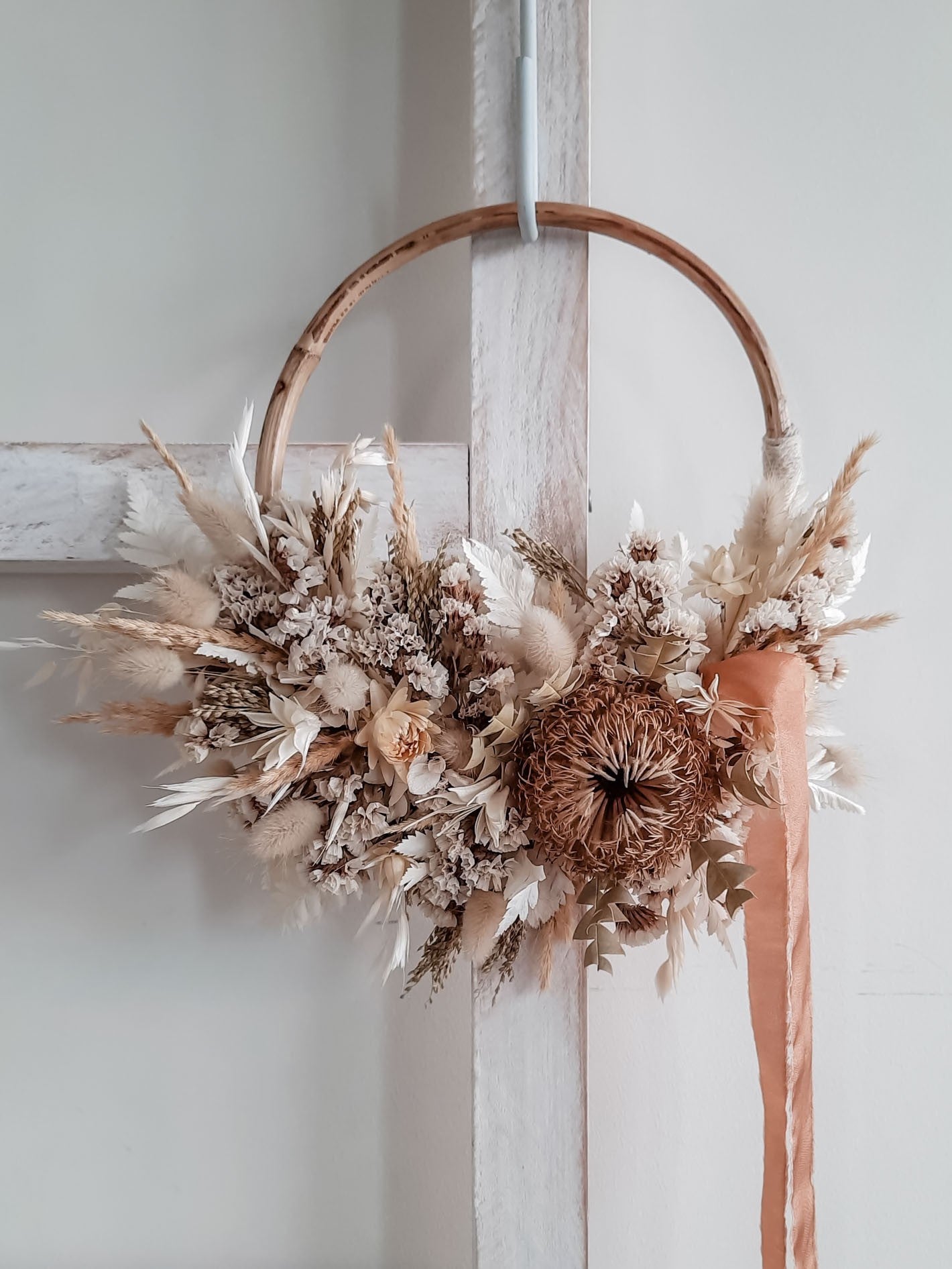 A rustic dried flower wreath featuring white and neutral dried flowers and foliage on a rattan hoop base, hanging on a white wooden ladder.
