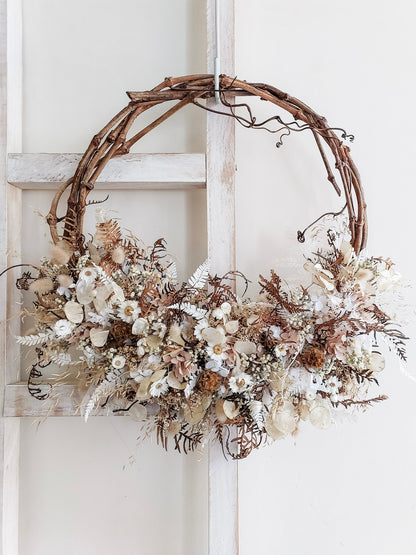 A rustic dried flower wreath featuring white, cream, and neutral dried flowers and foliage on a grapevine base, hanging on a white wooden ladder.