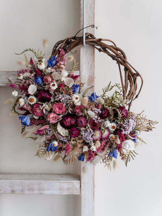 A rustic dried flower wreath featuring pink, plum, and blue dried flowers and foliage on a grapevine base, hanging on a white wooden ladder.