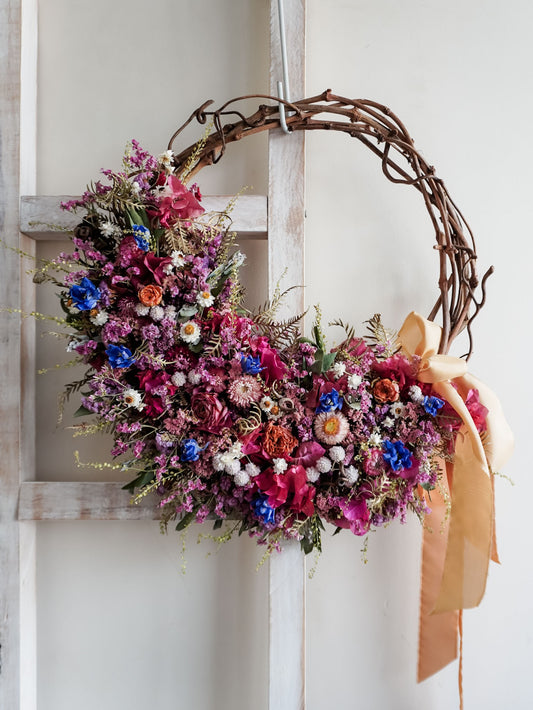 A rustic dried flower wreath featuring pink, blue, and purple dried flowers and foliage on a grapevine base, hanging on a white wooden ladder.