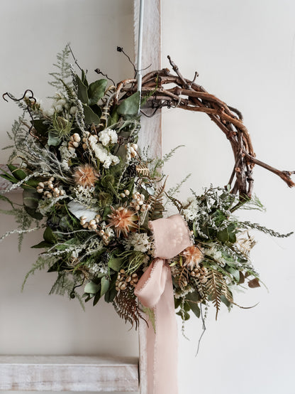 A rustic dried flower wreath featuring white, green, and earthy dried flowers and foliage on a grapevine base, hanging on a white wooden ladder.