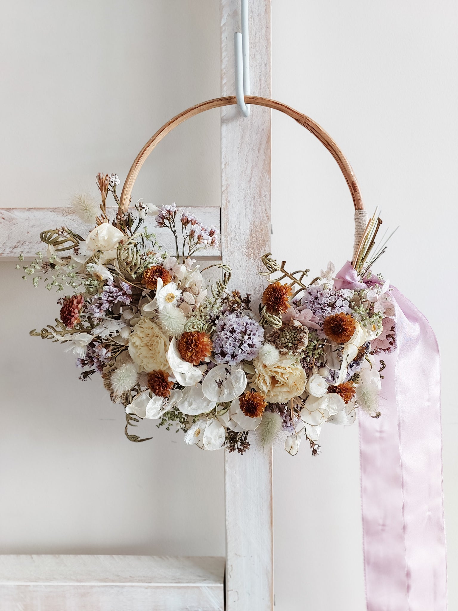A rustic dried flower wreath featuring mauve, cream, and rust dried flowers and foliage on a rattan hoop base, hanging on a white wooden ladder.