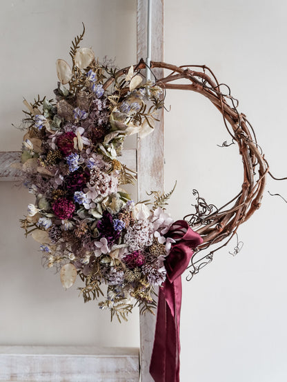 A rustic dried flower wreath featuring mauve, plum, and blue dried flowers and foliage on a grapevine base, hanging on a white wooden ladder.
