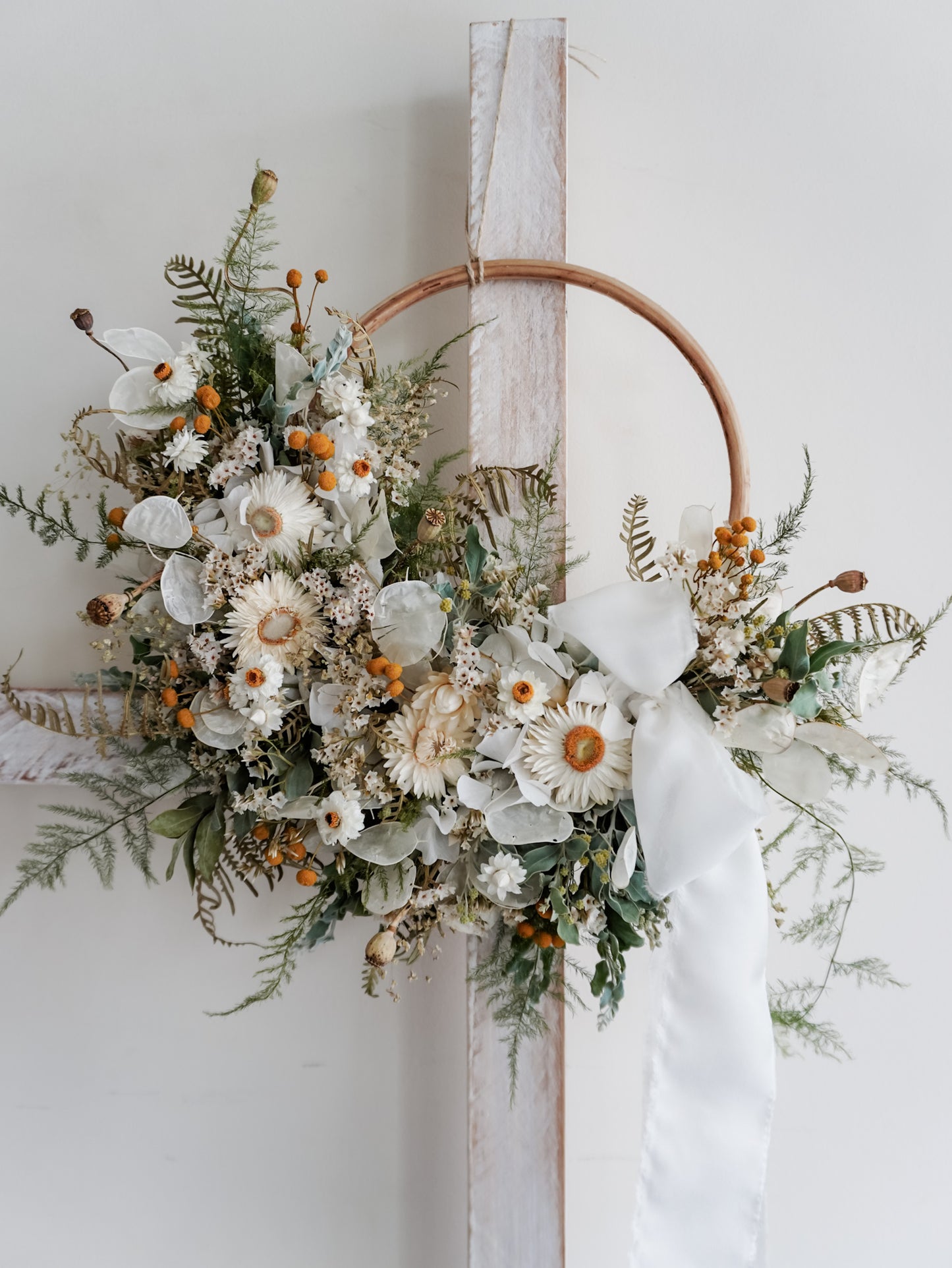 A rustic dried flower wreath featuring white, green, and yellow dried flowers and foliage on a rattan hoop base, hanging on a white wooden ladder.