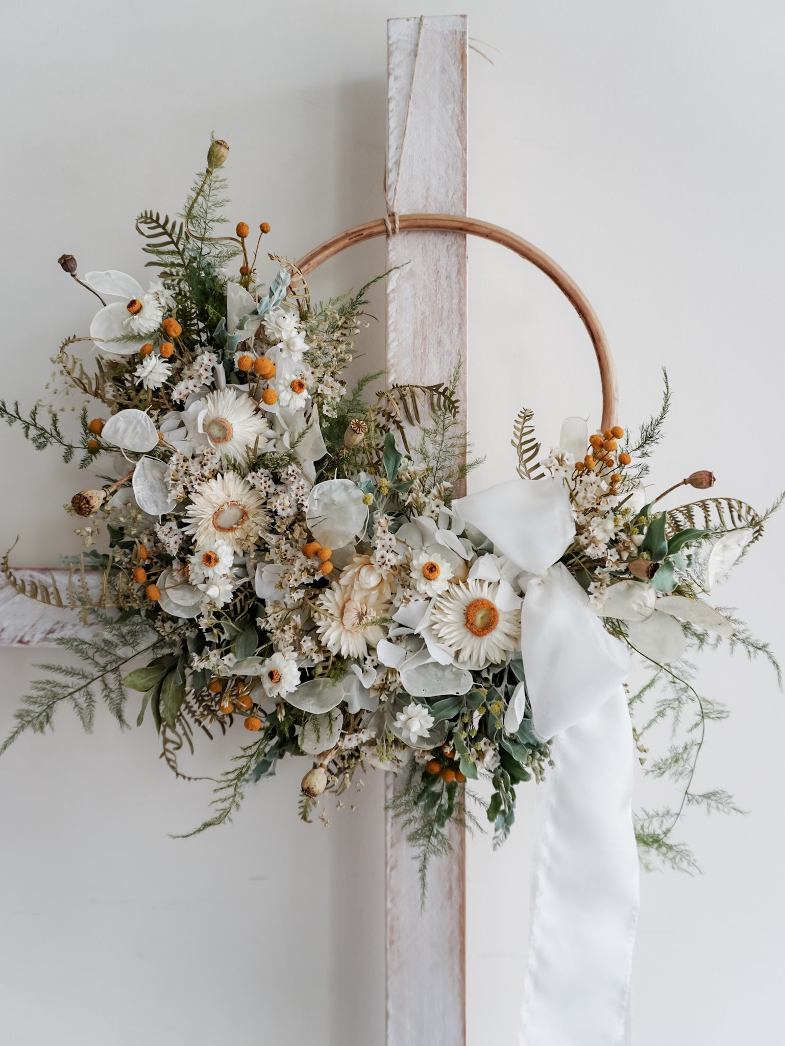 A rustic dried flower wreath featuring white, green, and yellow dried flowers and foliage on a rattan hoop base, hanging on a white wooden ladder.