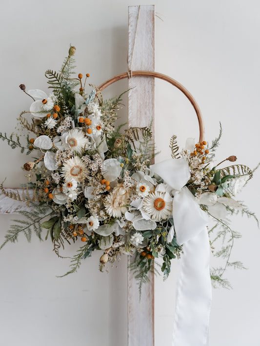 A rustic dried flower wreath featuring white, green, and yellow dried flowers and foliage on a rattan hoop base, hanging on a white wooden ladder.