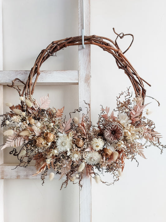 A rustic dried flower wreath featuring nude, cream, and dusty rose dried flowers and foliage on a grapevine base, hanging on a white wooden ladder.