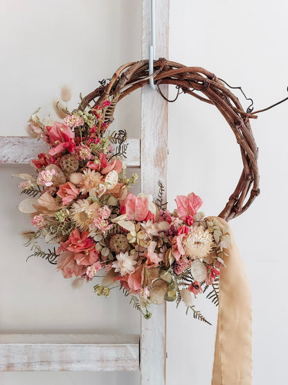A rustic dried flower wreath featuring pink, cream, and neutral dried flowers and foliage on a grapevine base, hanging on a white wooden ladder.