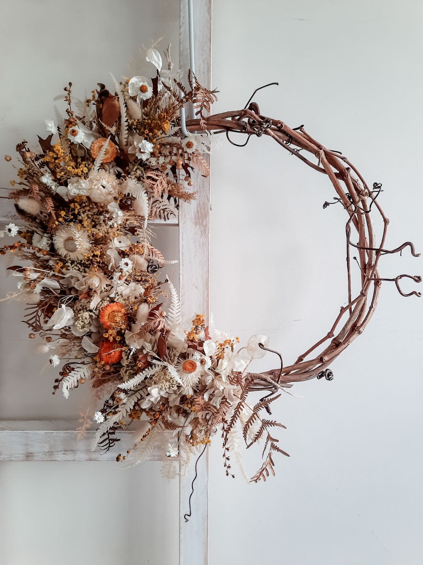 A rustic dried flower wreath featuring cream, gold, and earthy dried flowers and foliage on a grapevine base, hanging on a white wooden ladder.
