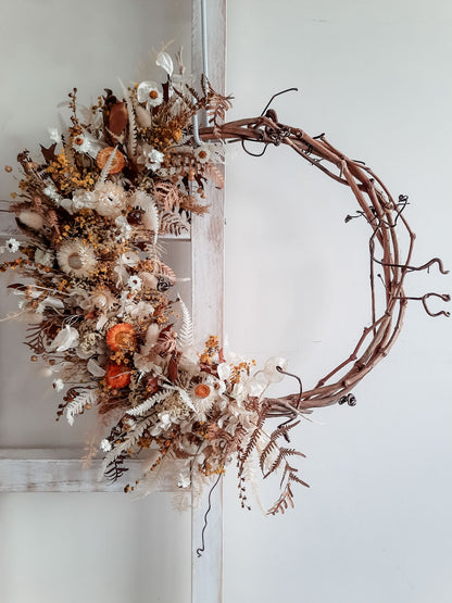 A rustic dried flower wreath featuring cream, gold, and earthy dried flowers and foliage on a grapevine base, hanging on a white wooden ladder.