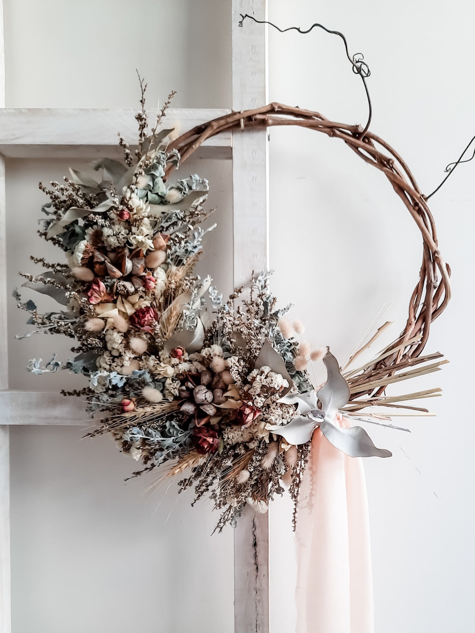 A rustic dried flower wreath featuring pastel, cream, and neutral dried flowers and foliage on a grapevine base, hanging on a white wooden ladder.