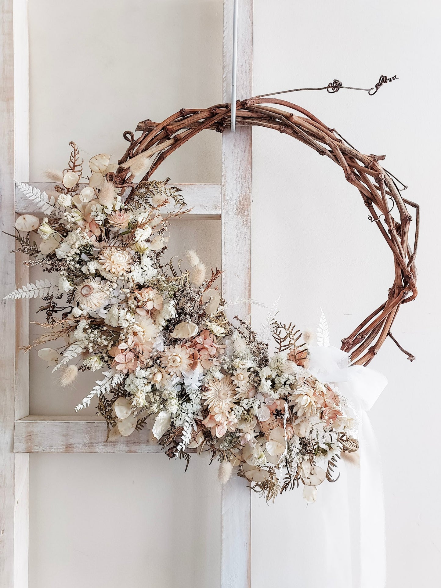 A rustic dried flower wreath featuring nude, cream, and white dried flowers and foliage on a grapevine base, hanging on a white wooden ladder.