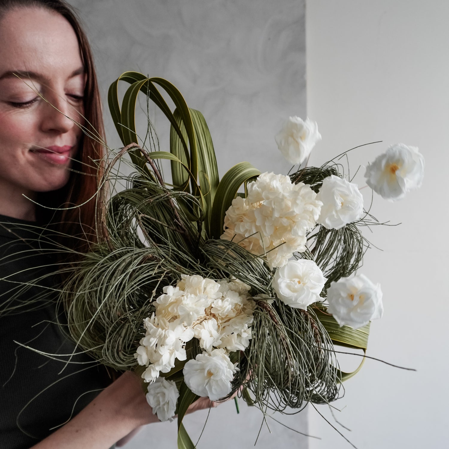 Florist holding a dried flower bridal bouquet of white flowers and greenery against a plain background